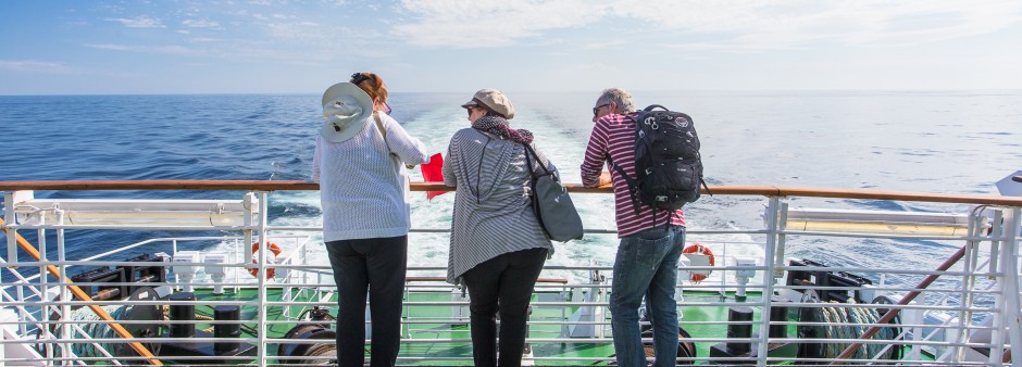 Trois personnes sont à l'arrière du traversier menant vers les Îles, ils regardent l'horizon. La mer est bleue, le temps est beau. Le soleil est au rendez-vous. C'est une belle traversée.