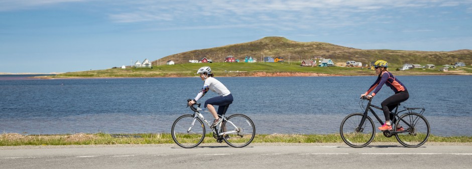 Two people cycle on the shoulder of a scenic road on the Îles de la Madeleine