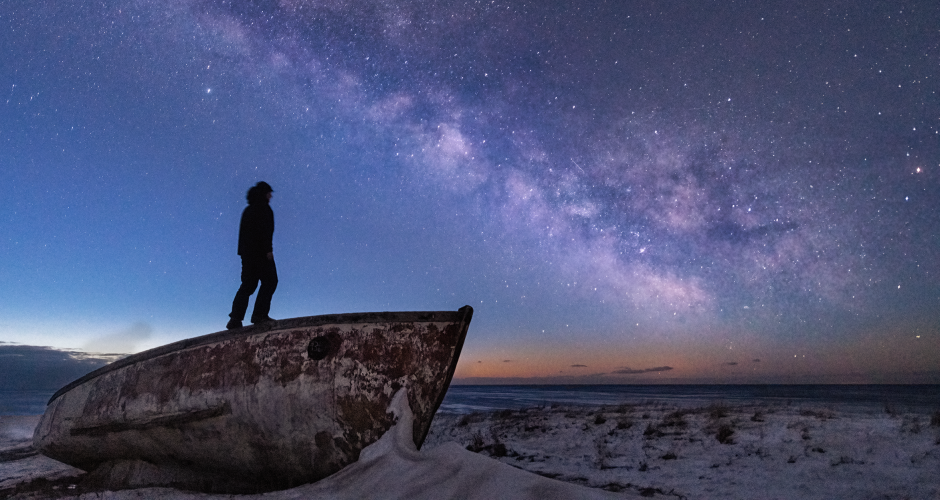 Un homme se tient debout sur une barque abandonnée sur un terrain aux Îles de la Madeleine. Il observe la voie lactée.